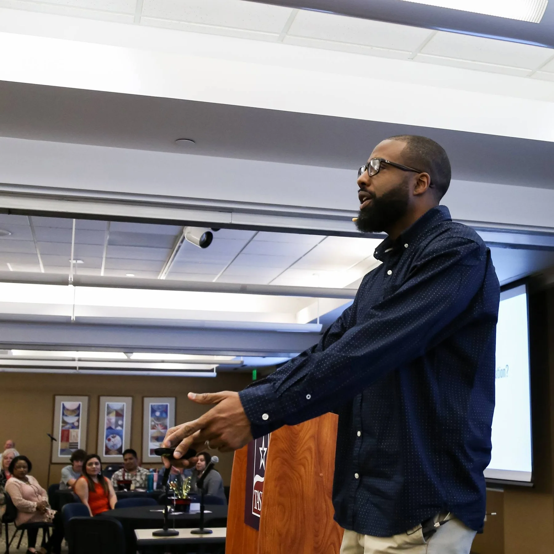 Bavu Blakes gesturing with both hands beside a wooden podium, with a seated audience behind him